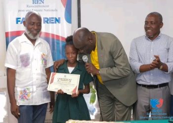 L’ancien ministre Jimmy Albert, le dos penché et un micro à la main, remet un certificat à une récipiendaire.